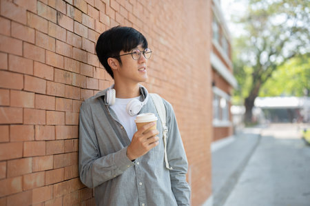 A smiling asian man with glasses is holding a coffee cup and looking a way while standing leaning against a brick wall. Take a rest or Chilling Outdoor.の写真素材