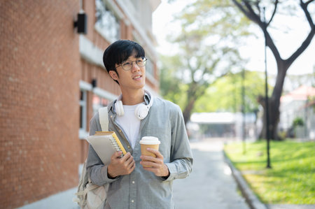 A smiling asian man with glasses is holding books with a coffee cup and looking away while standing near a brick wall. Walking outdoor or Going somewhere.の写真素材
