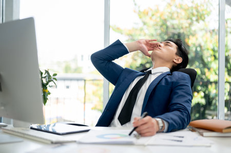 Asian businessman or male office worker is closing eyes and resting head on chair backrest to massaging his temples while sitting at working desk in office.の写真素材