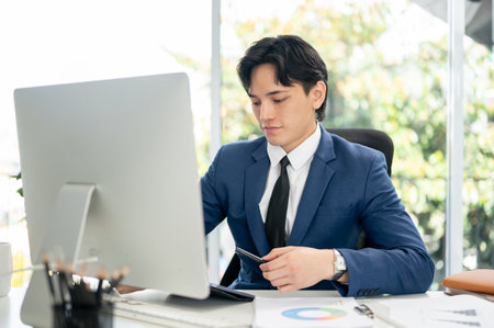 Businessman or male office worker in blue suit is holding a pen while looking down at his digital tablet on table. Sitting at working desk in office.の写真素材