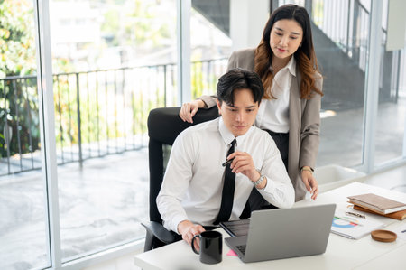 Asian female office worker and male coworker is looking at the data on laptop screen together. Businessman and woman is working in office.の写真素材