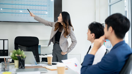 Female office worker is explaining and pointing at datas on monitor to her male coworkers. Businessman and businesswoman is having a meeting in office.の写真素材