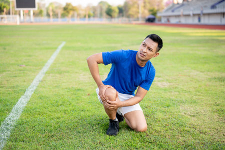Asian man in blue t shirt is kneeling and holding his knee while looking up. Leg pain or injury. Outdoor exercise or training. Sport and health.の写真素材