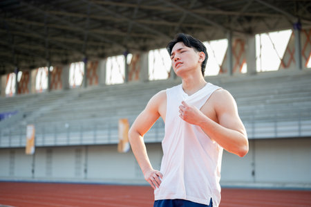Asian man standing with one hand on waist and other hand grabbing a white tank top, feeling hot or tired after running. Outdoor exercise or training.の写真素材