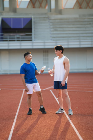 Two asian athlete men smiling at each other while holding their water bottles and standing on racetrack. Jogging or Running, Outdoor exercise or training.の写真素材