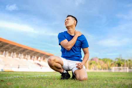 Asian man in blue t shirt is kneeling on grass and touching or massaging his shoulder. Pain or injury. Outdoor exercise or training. Sport and health.の写真素材