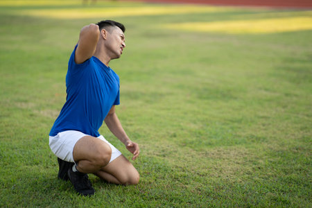 An asian man in blue t shirt touching his back while kneeling on the grass field. Back pain or injury. Outdoor exercise or training. Sport and health.の写真素材