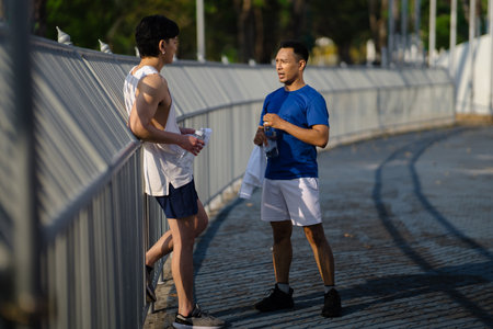 Two asian men or male friends are taking a rest and drinking water at sports stadium fence or railing. Outdoor exercise or work out, Athlete training.の写真素材