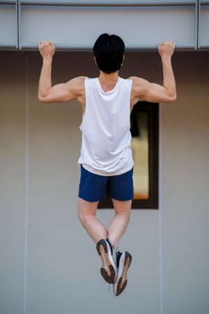 Full body of an asian man in white tank top doing a pull up on horizontal bar in sports stadium. Muscle Building, Outdoor exercise or or work out.の写真素材