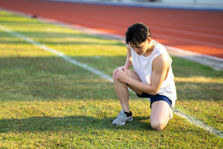 White tank top asian man with hands on his knee is crouching on grass field. Pain or injury. Outdoor exercise or training. Sport and health.の写真素材