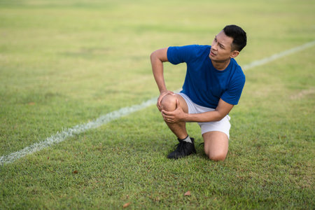 An asian man in blue t shirt is kneeling and holding his knee on the grass field. Knee pain or injury. Outdoor exercise or training. Sport and health.の写真素材