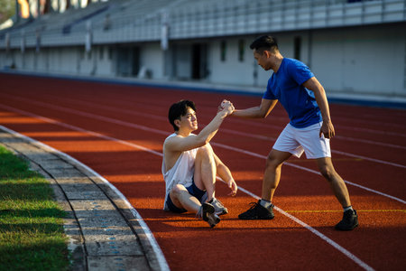 Asian man is grabbing or pulling his friend hand to help him standing up after falling on racetrack in sports stadium. Outdoor exercise or training.の写真素材