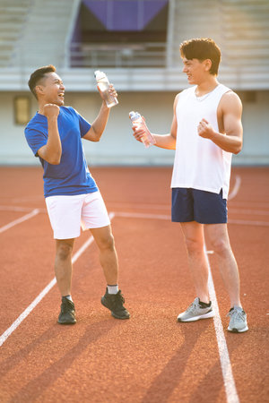 Two asian athlete men are holding their water bottles while talking together on the racetrack in sports stadium. Jogging or Running, Outdoor exercise.の写真素材
