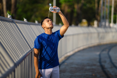 Asian man in blue t shirt is pouring water from his water bottle on himself. Reducing the heat or cooling down. Outdoor exercise or work out.の写真素材