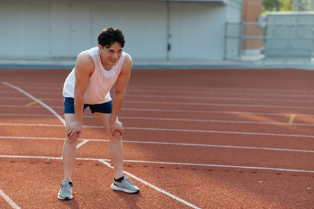 Asian man in white tank top looking ahead while standing with hand on his knee while take a rest after run on racetrack in sports stadium. Outdoor exercise.の写真素材