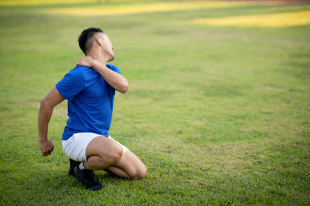 An asian man in blue t shirt is touching his shoulder and turning his head while sitting on grass field. Pain or injury. Outdoor exercise. Sport and health.の写真素材