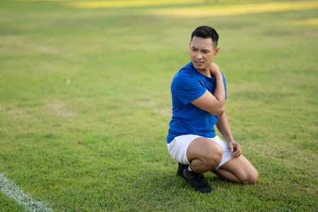An asian man in blue t shirt is touching his shoulder and looking away while kneeling on grass field. Pain or injury. Outdoor exercise. Sport and health.の写真素材