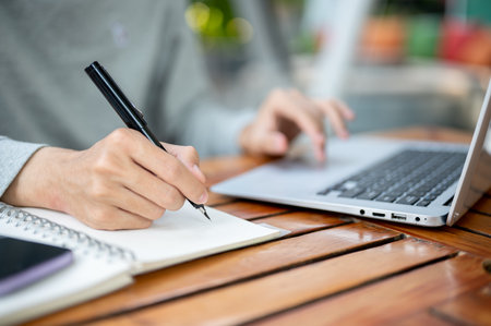 Close up of a hand holding a pen writing in the notebook and other hand tapping on laptop while sitting at wooden table. Study or working outdoor.の写真素材