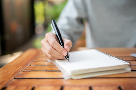 Close up of a hand holding a pen writing in the notebook while sitting at wooden table in the cafe or coffee shop. Study or working outdoor.の写真素材