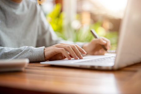 Close up of a hand holding a pen and other hand touching on laptop while sitting at wooden table in a cafe or coffee shop. Study or working outdoor.の写真素材