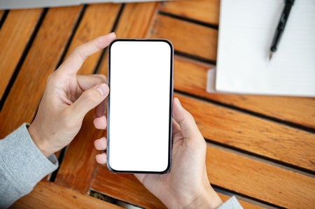 Top view of hands holding a blank screen or white screen smartphone on wooden table. Empty screen mock up for product display or advertisement.の写真素材