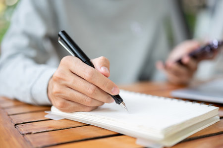 Close up of a hand holding a pen writing in the notebook and other hand holding a smartphone while sitting at wooden table. Study or working outdoor.の写真素材