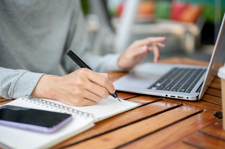 Close up of a hand holding a pen writing in the notebook and other hand hovering over laptop while sitting at wooden table. Study or working outdoor.の写真素材