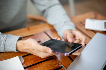 Close up of a hand holding and using a smartphone on wooden table while sitting in the coffee shop or cafe. Study or working outdoor.の写真素材