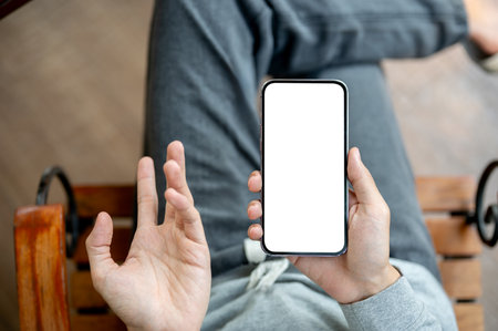 One hand holding white screen smartphone with other hand in an explaining gesture, sitting cross legged on chair. Empty screen mock up, Product display, Advertisement.の写真素材