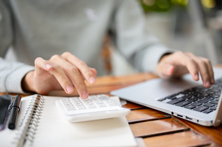 Close up of a hand using white calculator on the notebook and other hand resting on laptop while sitting at wooden table. Study or working outdoor.の写真素材