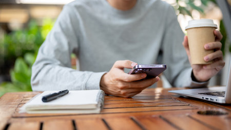A man holding a smartphone and other hand holding a coffee cup while sitting at wooden table in the cafe or coffee shop. Study or working outdoor.の写真素材
