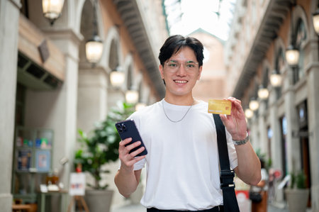 Asian man with glasses in white tee is smiling while holding phone and showing his credit card in...