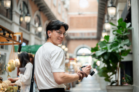 Asian man wearing glasses in white tee is looking back while holding a camera in the hallway of...