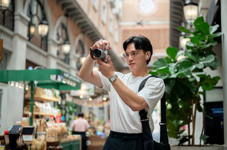 Asian man wearing glasses in white tee is taking a photo on his camera in the hallway of shopping...