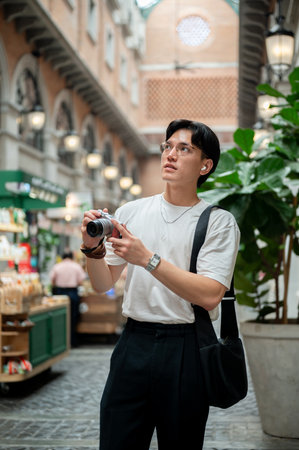 Asian man wearing glasses in white tee is looking up while holding a camera in the hallway of shopping mall or indoor market. Photo trip. Tourist spot.の写真素材