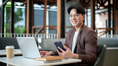 Brown suit asian man with glasses is smiling while holding his smartphone, sitting with laptop in...
