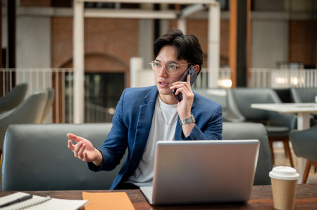 Blue suit asian man is talking on the phone with explaining hand gesture while sitting with a laptop in cafe or coffee shop. Study or working outside.の写真素材