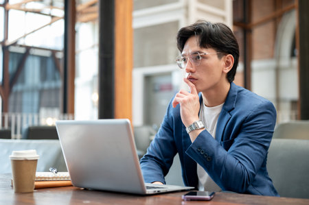 Blue suit asian man is thinking thoughtfully in front of his laptop while sitting at wooden table in cafe or coffee shop. Study or working outside.の写真素材