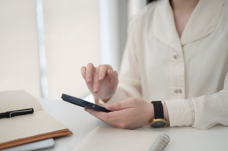 Close up of female office worker hand hovering over smartphone while sitting at working table in office. Desk Job, Corporate Job, Modern Company.の写真素材