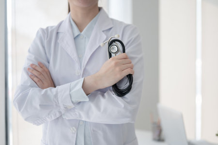 Close up of a female doctor standing with arms crossed while holding a black stethoscope in the diagnosis room, Hospital or Clinic, Doctor's office.の写真素材