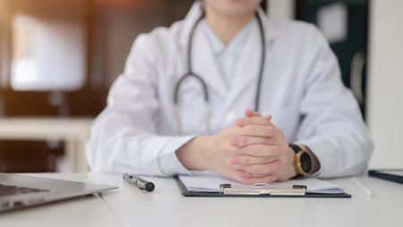 A doctor with stethoscope clasping hands over documents on clipboard, sitting at white table in the diagnosis room, Hospital or Clinic office.の写真素材