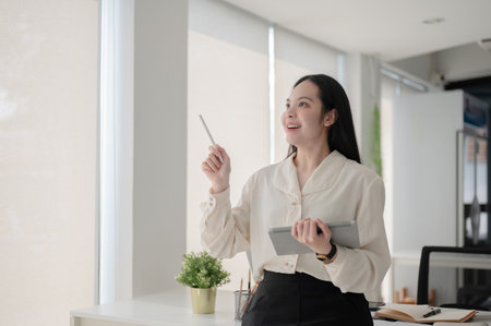 Female office worker smiling and looking away from tablet while standing in front of working table....