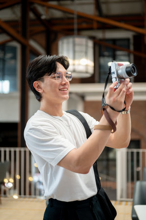 Asian man wearing glasses in white T shirt is smiling while holding his camera taking a photo in the cafe or coffee shop. Photo trip, Chilling outside.の写真素材