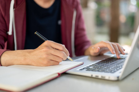 Close up of a hand holding ballpoint pen writing in notebook and another hand typing on laptop while...