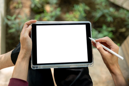 Top view of a hand holding white screen tablet and stylus pen while sitting cross legged near plant...