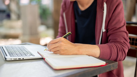 Side view of a hand writing in notebook with a ballpoint pen while sitting in front of a laptop at...
