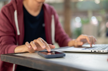Close up of a hand touching on smartphone with another hand typing on laptop while sitting at...