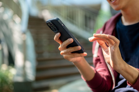 Close up of an asian man looking and tapping on his smartphone, watching videos or making a call while sitting near the stair. Chilling Outdoor.の写真素材
