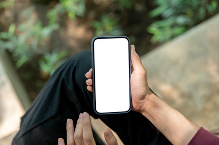 Close up of a hand holding a white screen smartphone vertically while sitting cross legged near the plant bed. Looking or Watching, Chilling Outdoor.の写真素材
