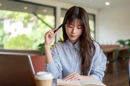 Woman in blue striped shirt is thinking and holding a fountain pen while looking at the notebook sitting in the cafe. Study or Working Outside.の写真素材